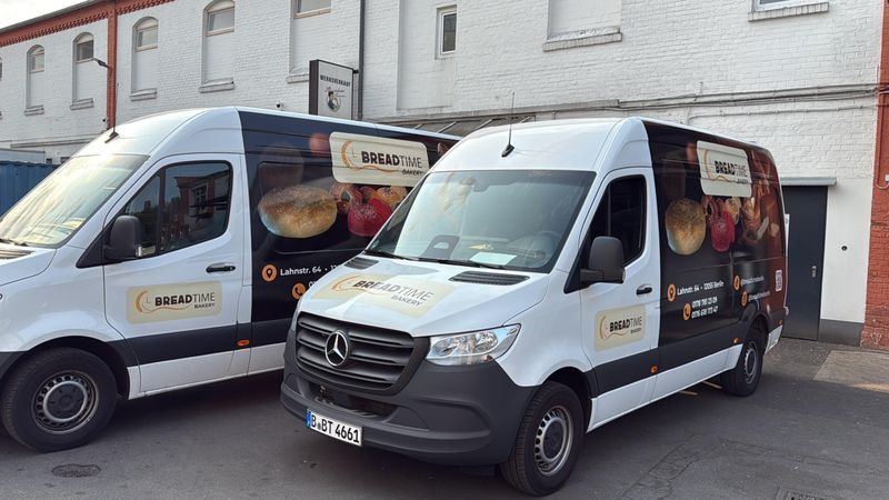 Zwei Lieferfahrzeuge von Breadtime Bakery nebeneinander auf einem Hof, beklebt mit dem Logo und Abbildungen von Backwaren